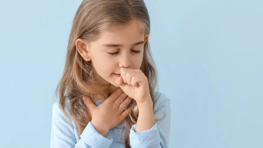 Uma menina com uma mão no peito e a outra perto da boca, tossindo ou com dor de garganta, ilustrando sintomas de resfriado em crianças.
